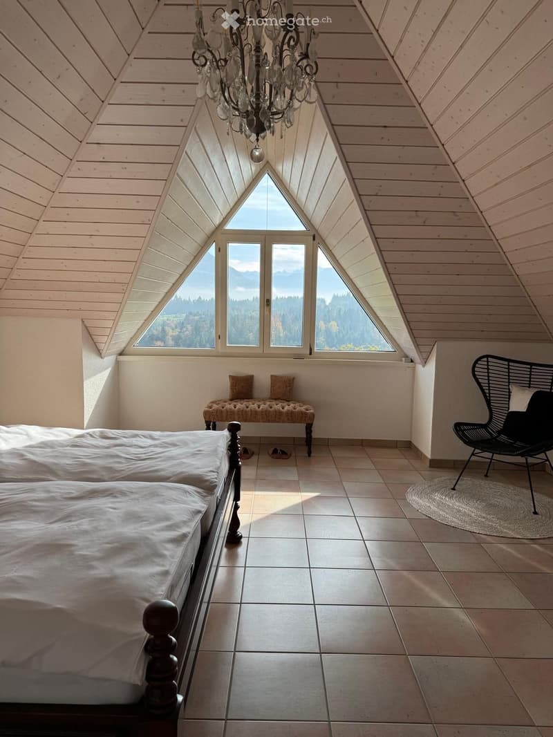 Bright, airy bedroom with a slanted wooden ceiling, large windows offering a scenic view, a double bed, a bench, a modern chair, and a round rug.