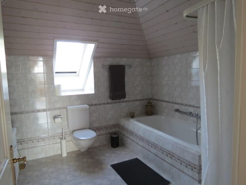 Bathroom with a toilet, bathtub, towel, shower curtain, and a small window. The walls are tiled, and there is a rug on the floor.