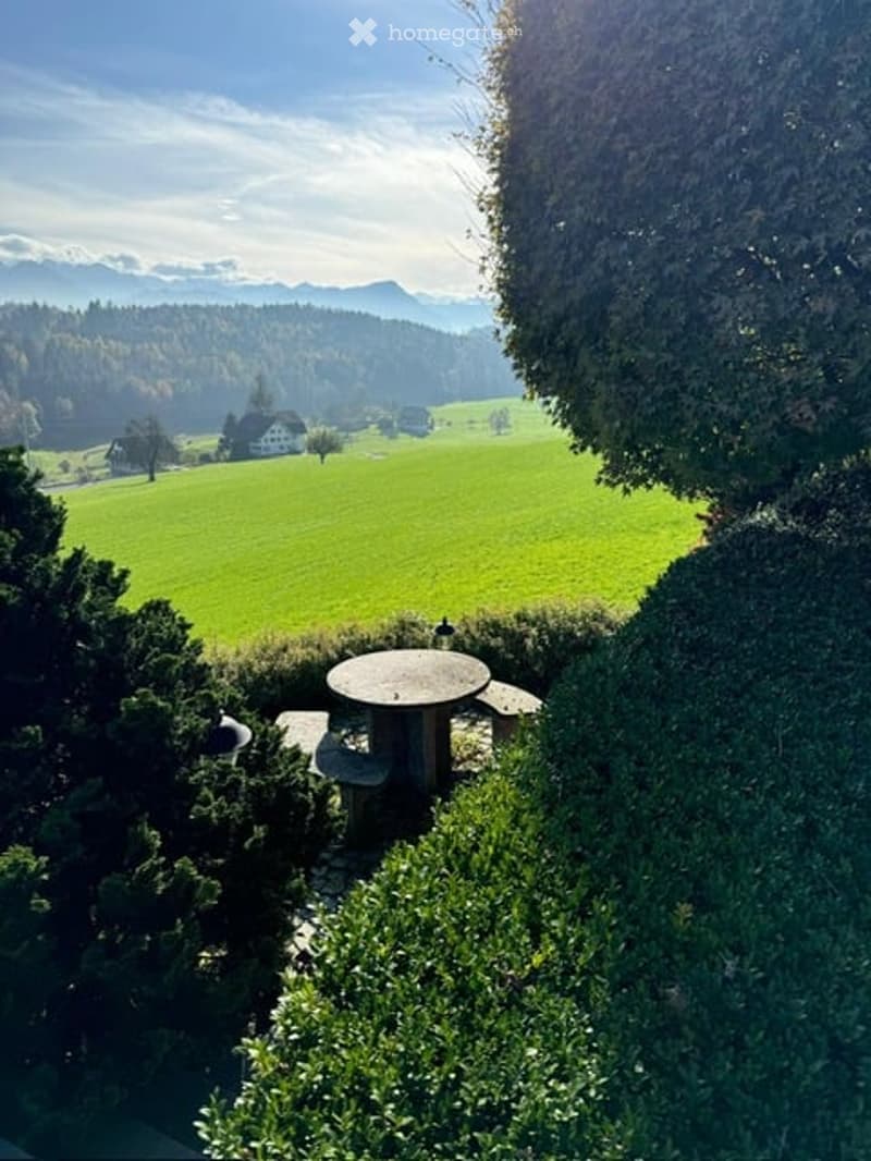 Outdoor seating area with a round table and benches surrounded by greenery, overlooking a green field and distant mountains.