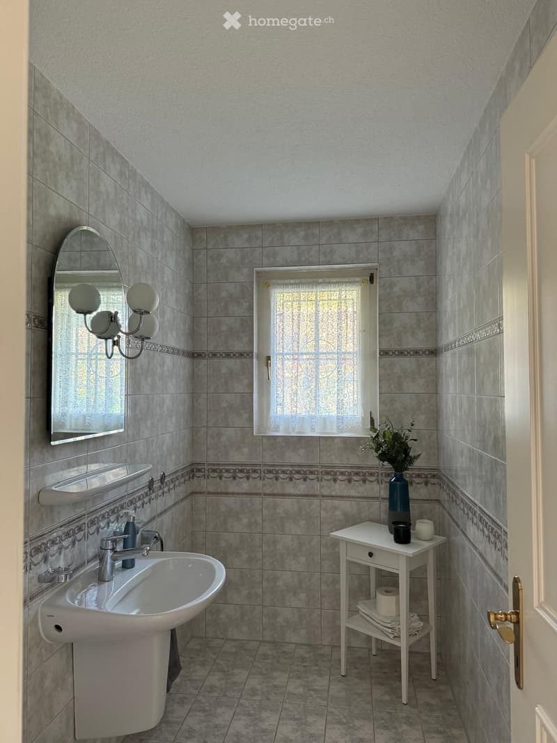 Bathroom with a sink, mirror, small table with vase and cup, and a window with a curtain.