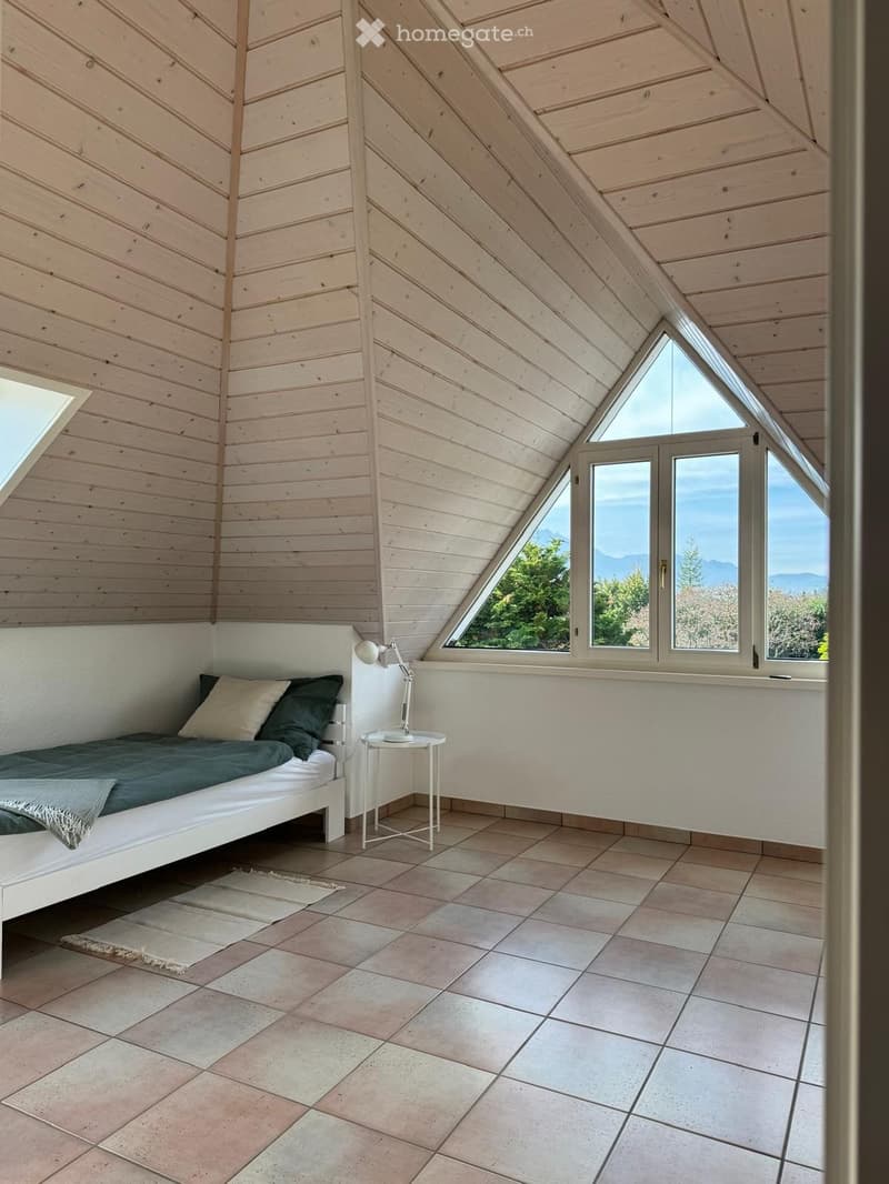Bright, modern bedroom with wooden walls and slanted ceiling, featuring a white bed, nightstand, and large windows offering a scenic view.