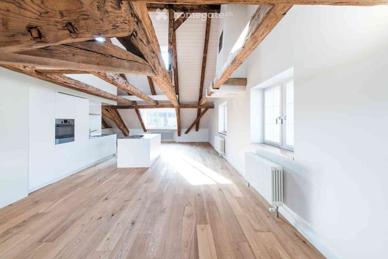 Bright, open room with exposed wooden beams, white walls, wooden flooring, and large windows allowing natural light. Kitchen area with white cabinets and appliances is visible on the left side.