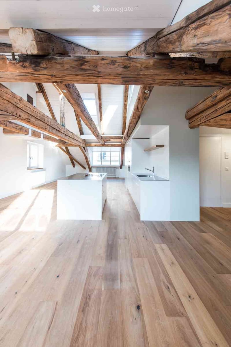 Bright, airy kitchen with exposed wooden beams, white cabinetry, and a central island. Light wooden flooring and large windows provide natural light.