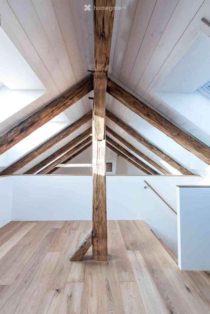 Empty attic space with exposed wooden beams and light wooden flooring, featuring a central wooden post and a staircase leading upwards.