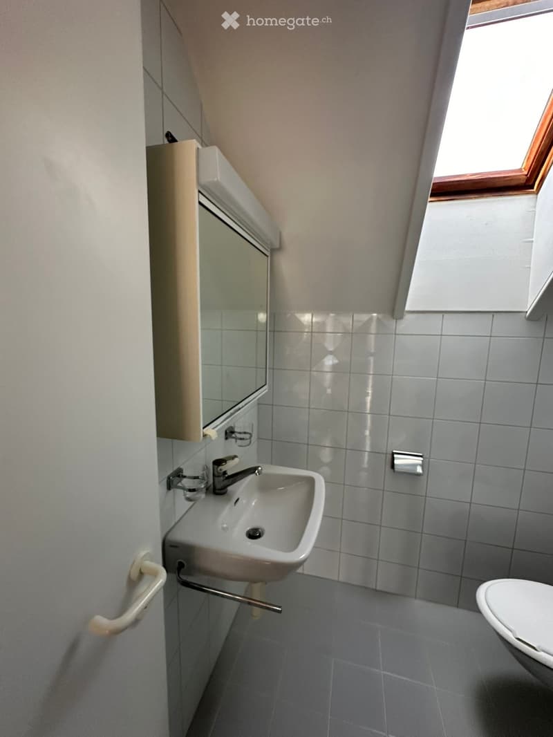 Small bathroom with a white sink, mirror, and toilet. Grey tiled walls and floor, window with wooden frame, and a cabinet with mirrored front.