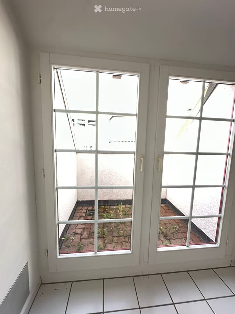 Empty room with white tiled floor, white walls, and large glass doors leading to a small outdoor area with a brick-paved surface and small plants.
