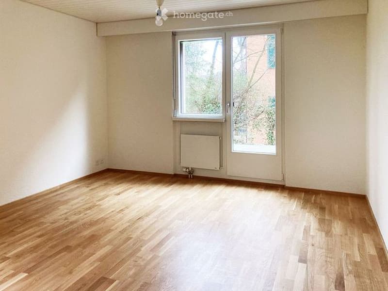 Empty room with wooden flooring, white walls, large window with blinds, radiator, and a door leading outside.