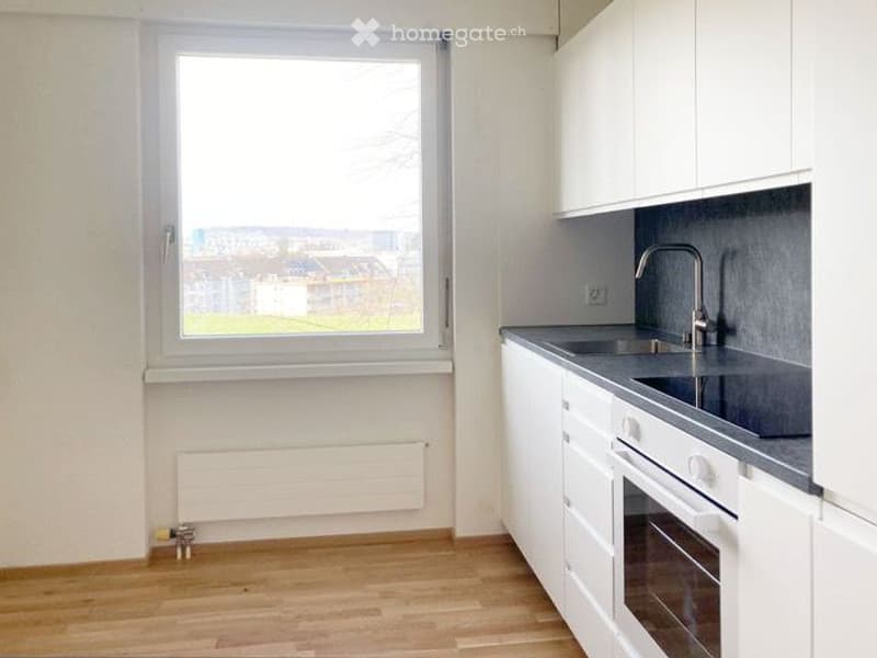 Modern kitchen with white cabinets, grey countertops, a sink with a faucet, and an oven. The room features wooden flooring and a large window offering a view of the outside.