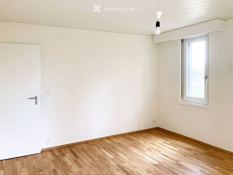 Empty room with light wooden flooring, white walls, a single window, and a closed white door.