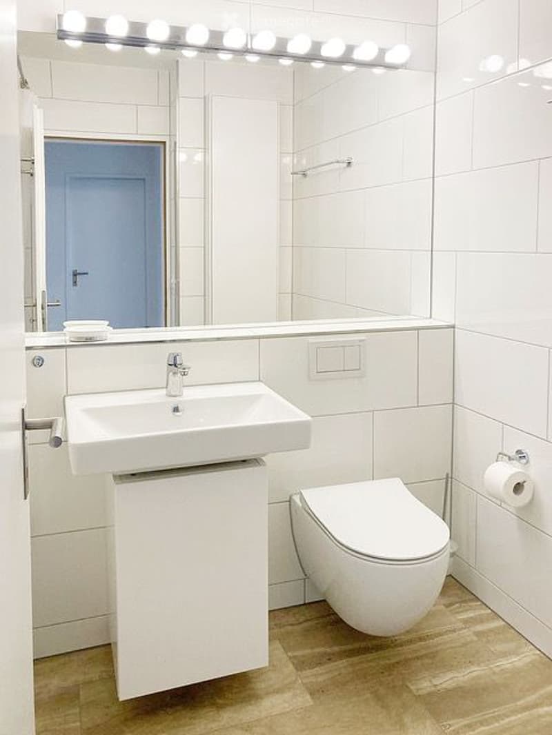 Modern bathroom with white tiles, a white sink, toilet, and a large mirror with integrated lighting above the sink.