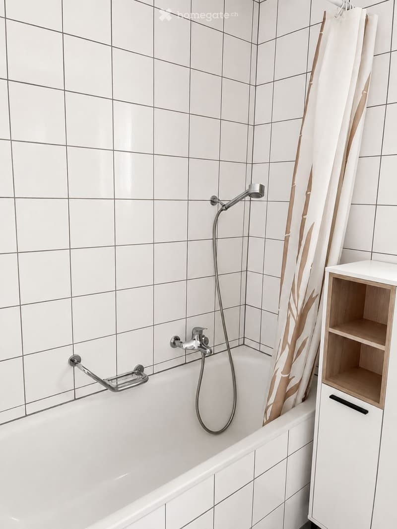 Modern bathroom with a white tiled wall, a bathtub, a handheld showerhead, a shower curtain, and a wooden cabinet with shelves.