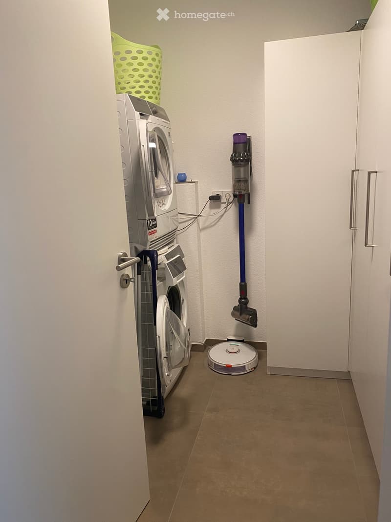 Small bathroom with a washing machine, a vacuum cleaner on a wall mount, and a robotic vacuum on the floor.
