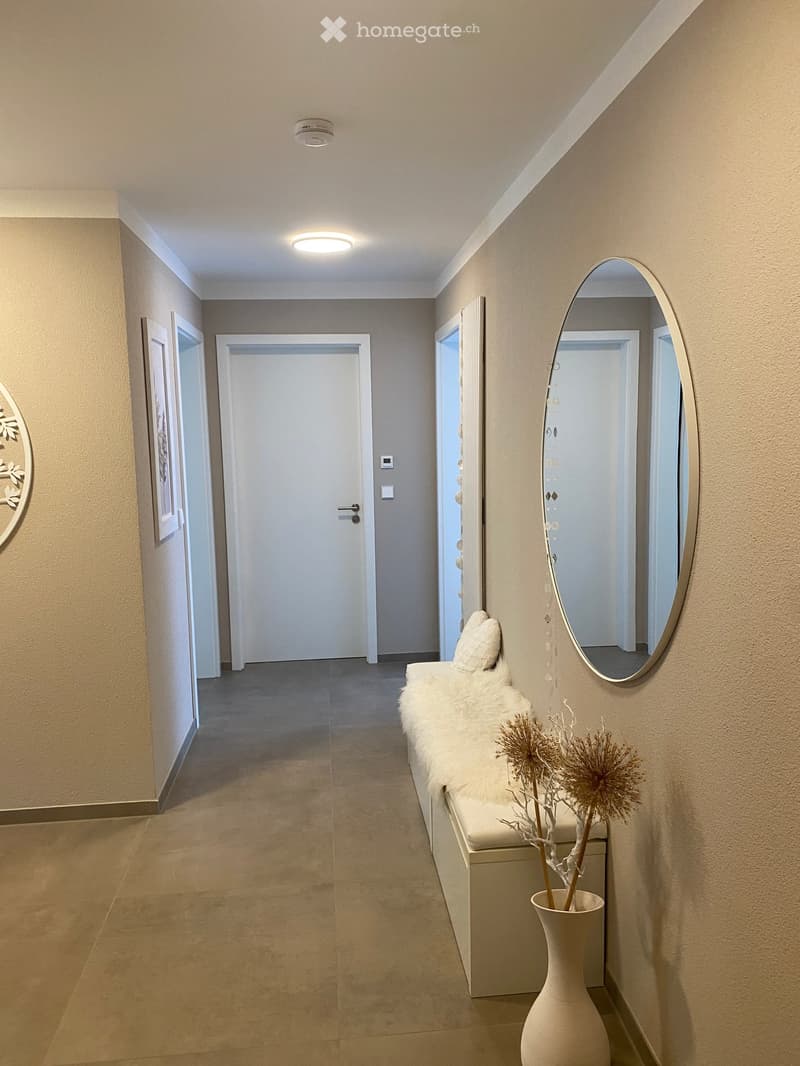 Modern hallway with beige tiled floor, white walls, recessed lighting, and a bench with a fur throw and decorative branches in a vase.