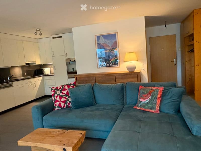 A modern living room with a blue sectional sofa, wooden coffee table, table lamp, and a small kitchen area with white cabinets and a microwave.