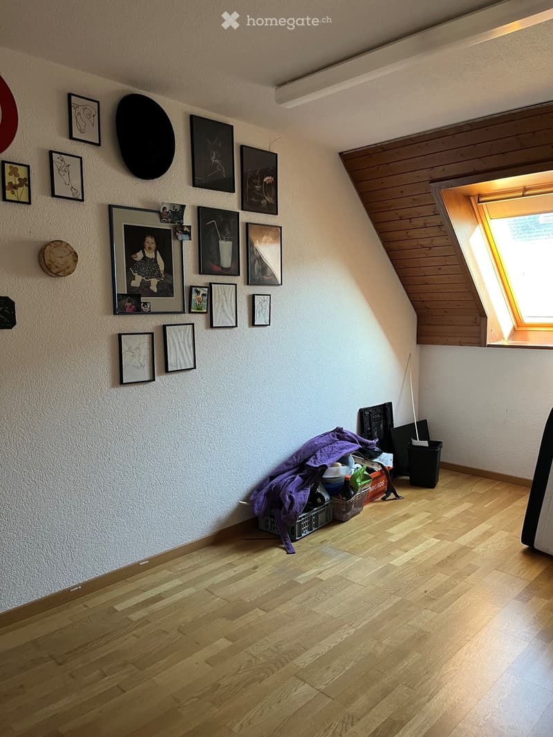 Empty room with wooden flooring and white walls, featuring a slanted ceiling and skylight window. Various framed pictures and artwork are displayed on the walls, and a small pile of items including a purple blanket and cleaning supplies is placed near the