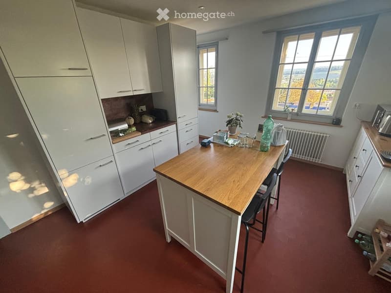 Modern kitchen with white cabinets, a central wooden island, and a red floor. The island has a wooden countertop and is surrounded by chairs. There are various items on the island, including a bottle, a flower pot, and other small objects.
