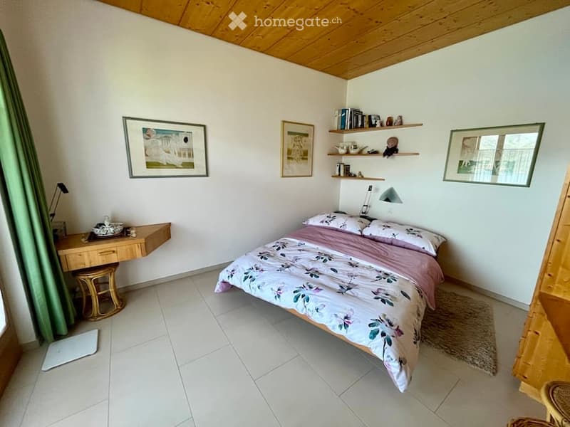 A cozy bedroom with a bed, a desk, a rug, and picture frames on the wall. Wooden ceiling and floor.