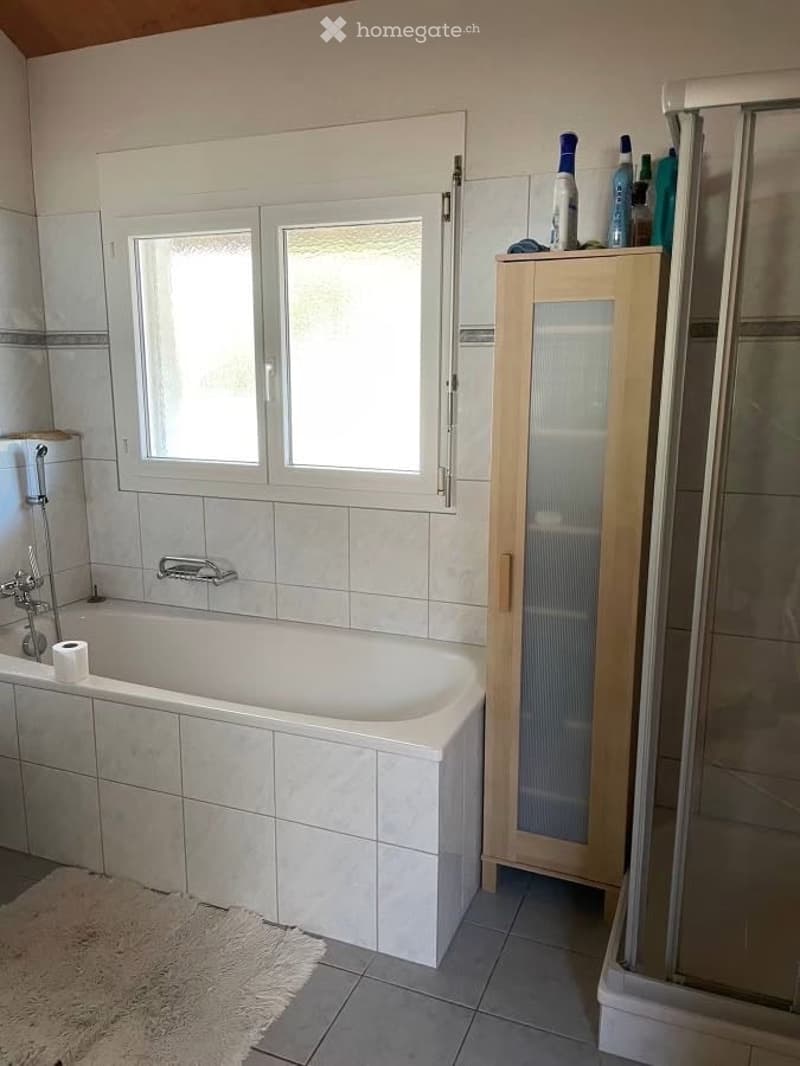 Bathroom with a white bathtub, tiled walls, a shower area, a wooden cabinet with glass doors, and several shampoo bottles on top. There is a window above the bathtub and a toilet paper roll on the edge of the tub.
