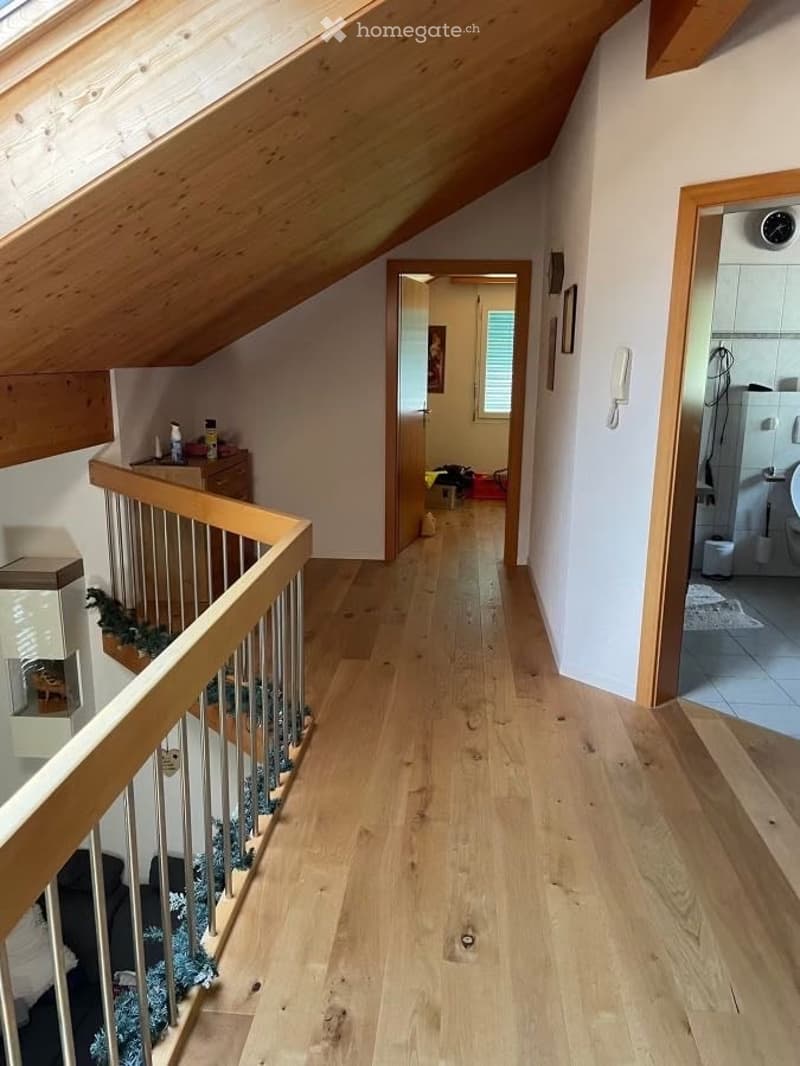 Spacious hallway with wooden flooring and a wooden staircase leading to an upper level. The hallway features a small cabinet with decorative items, a wall-mounted phone, and a view into a bathroom and another room.