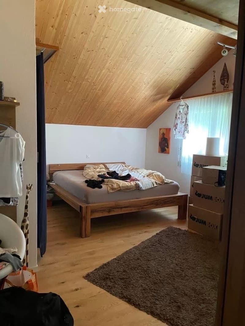 Cozy attic bedroom with wooden slanted ceilings, a bed with a wooden frame, a brown rug, and a wardrobe with clothes hanging. There are also some boxes and a picture frame on the wall.