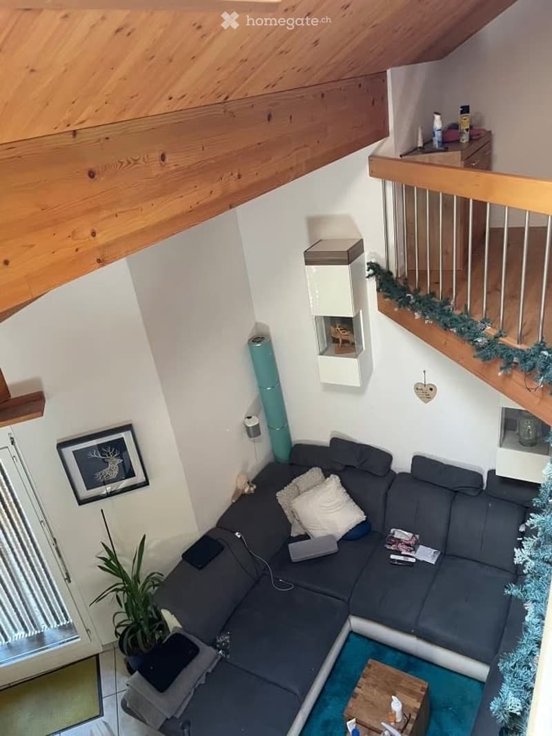 Modern living room with a grey sectional sofa, white throw pillows, a wooden coffee table, a potted plant, and a picture frame on the wall. The room features exposed wooden beams on the ceiling and a staircase leading to an upper level.