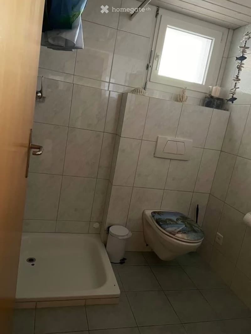 A small bathroom with a bathtub, toilet, and a window. The walls are tiled, and there is a trash bin near the bathtub.