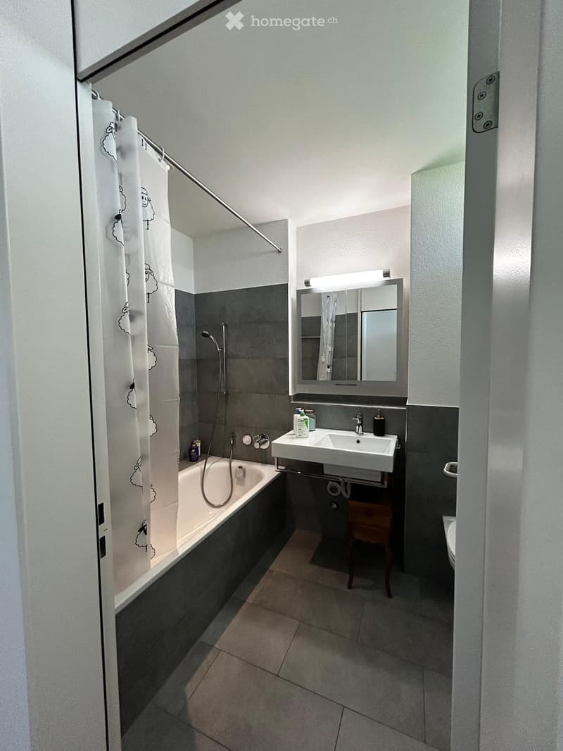 Modern bathroom with a bathtub, sink, mirror, and toilet. Features a shower curtain with a sheep design, a built-in shelf, and a small wooden stool.