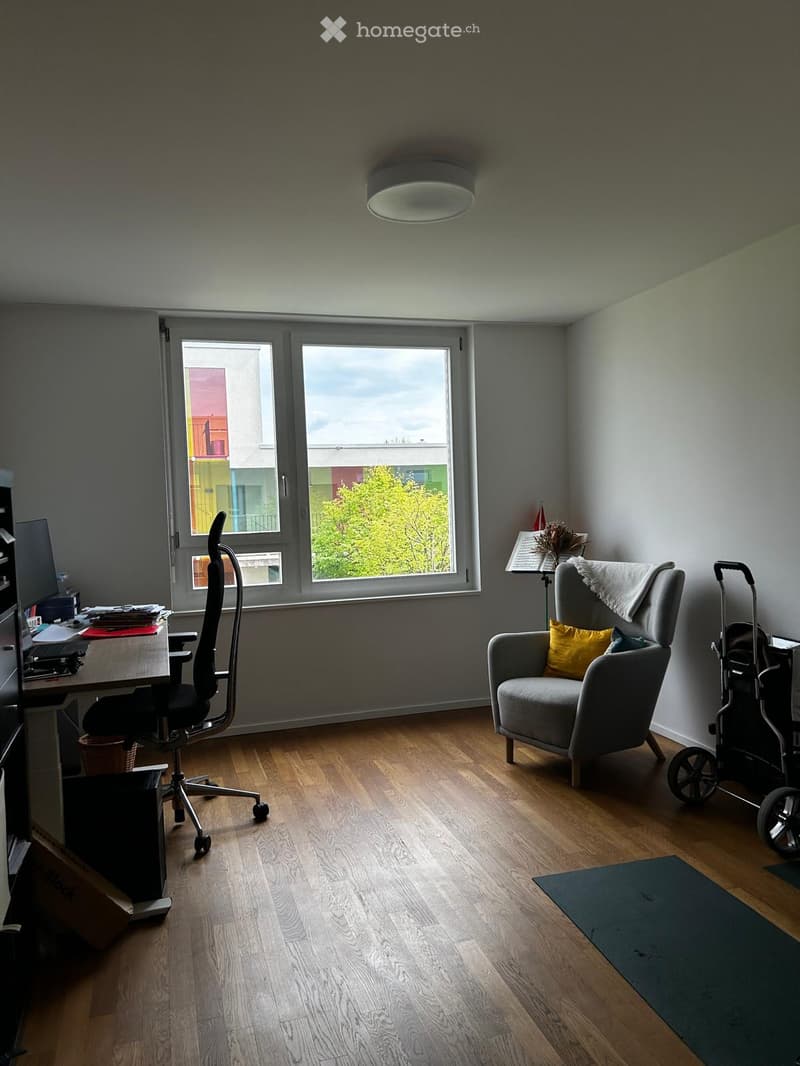 A modern, well-lit room with wooden flooring, large windows, a desk with a chair, a sofa with a pillow, and a small table with a lamp.