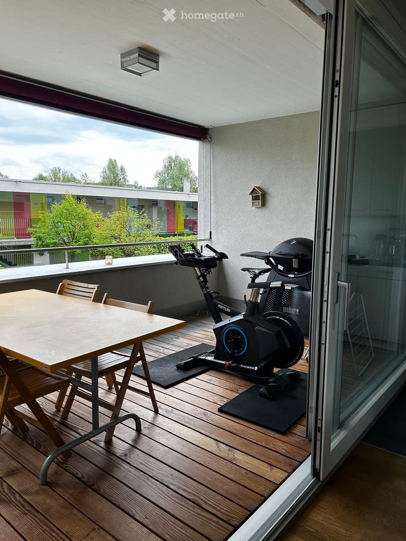 Balcony with wooden decking, a small table with chairs, and an exercise bike. The balcony has a glass door leading inside and a railing with a view of the surrounding area.