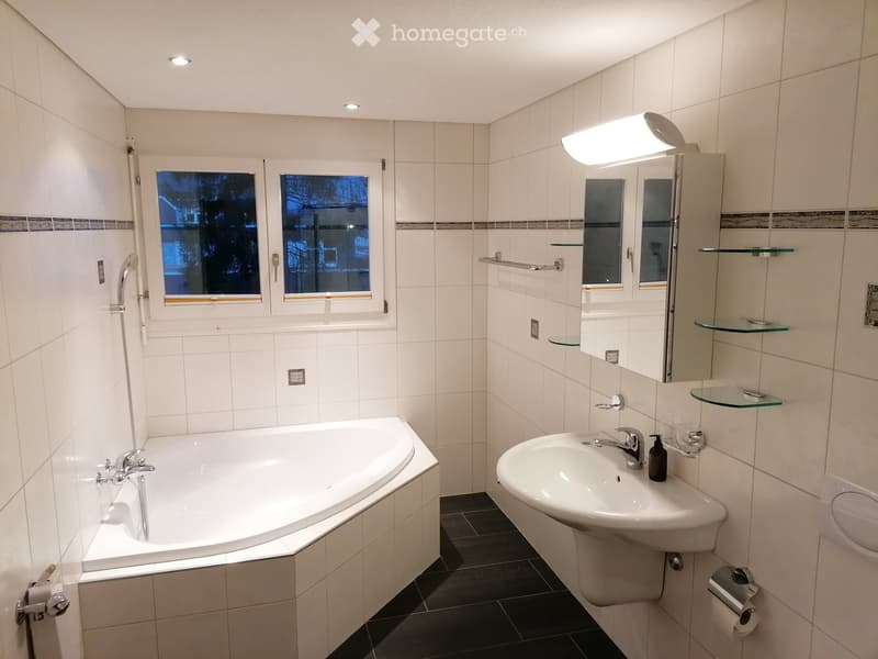 Modern bathroom with white tiled walls, a bathtub, a sink with a mirror, and glass shelves. The room features a window with a view of trees and a night sky.