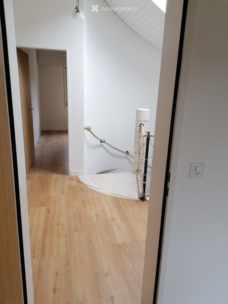 Modern hallway with light wooden flooring, white walls, and a decorative rope railing leading to an open area.
