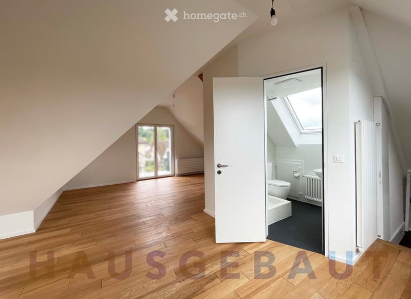 An unfurnished attic room with wooden flooring, slanted ceilings, and a door leading to a small bathroom. The bathroom features a toilet, sink, and a small window with a skylight above.