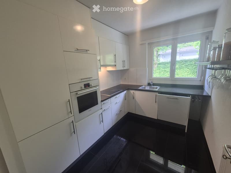 Modern kitchen with white cabinets, black countertops, and a built-in oven. Features a large window with a view of greenery.
