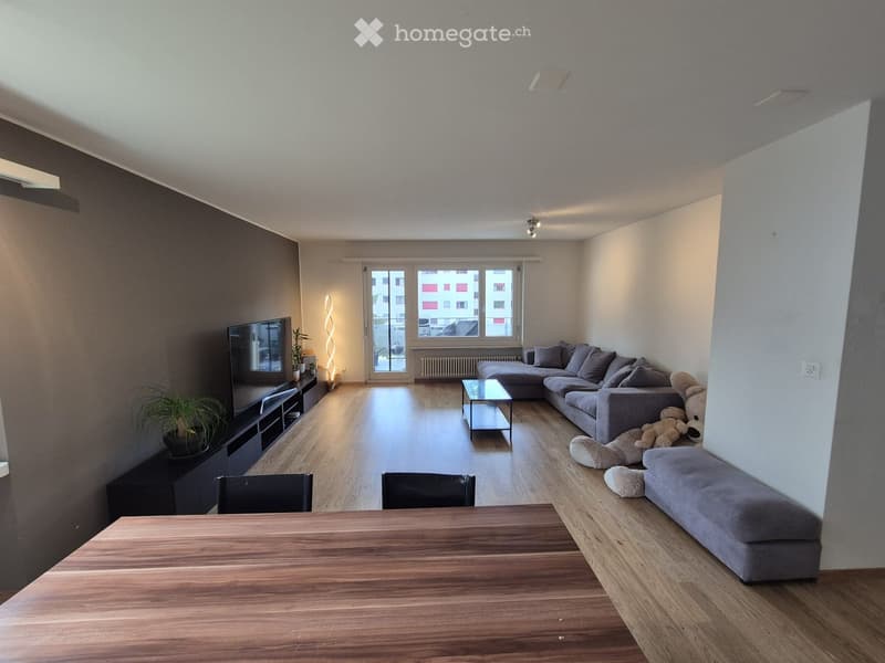 Modern living room with a grey sectional sofa, coffee table, dining table, chairs, TV, and decorative plants.