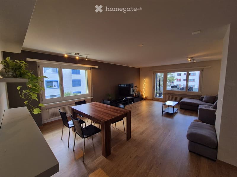 Open-plan living and dining area with wooden flooring, large windows, a sofa, ottoman, coffee table, dining table with chairs, and a TV stand with a television.