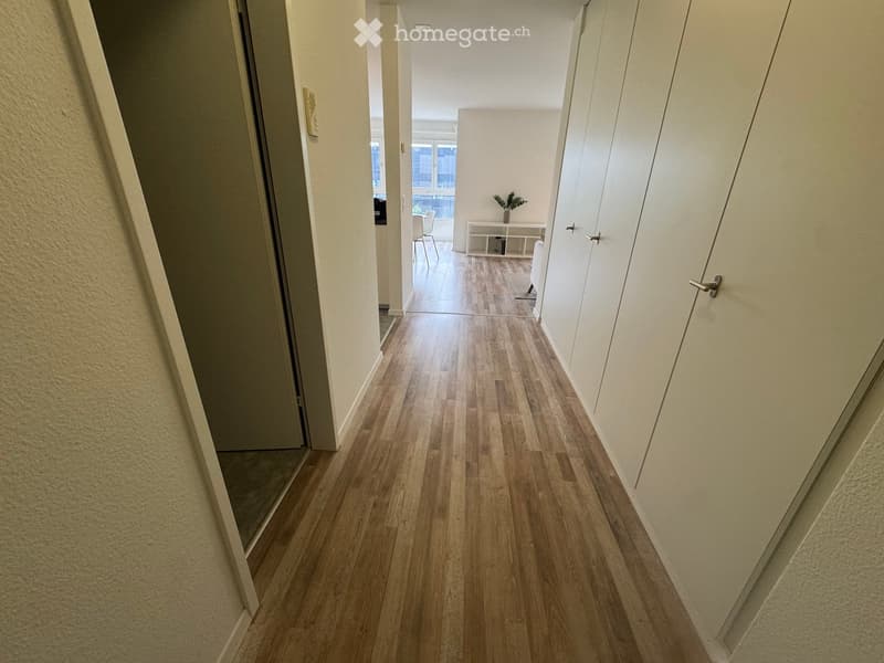 A clean, well-lit hallway with wooden flooring, white walls, and several doors leading to different rooms.