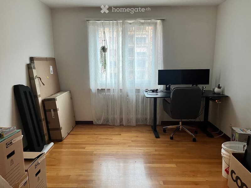 A small, furnished room with a wooden floor, a desk with a monitor and chair, and a window with white curtains. There are several cardboard boxes and a potted plant visible.