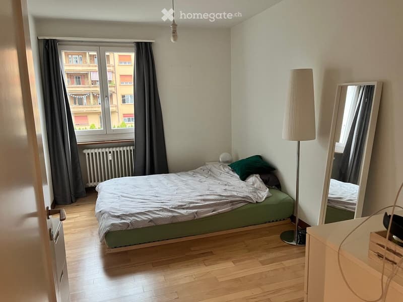 Cozy bedroom with wooden flooring, a neatly made bed with green bedding, a tall floor lamp, a large mirror, and a radiator beneath a large window with dark curtains.