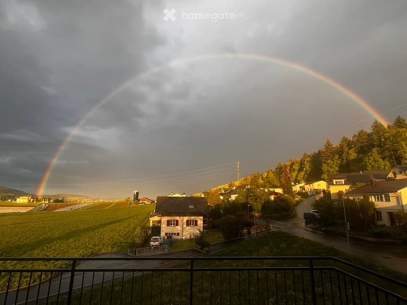 A scenic view from a balcony showing a rainbow over a rural landscape with houses, fields, and trees.
