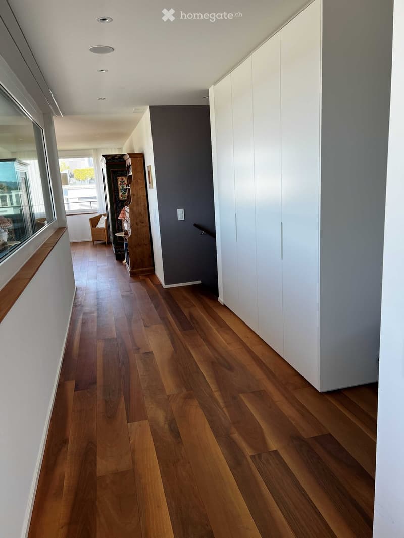 A modern hallway with wooden flooring, white walls, and built-in white cabinets. A small seating area with a wicker chair and a small table is visible in the background, along with a large window offering a view of the outside.