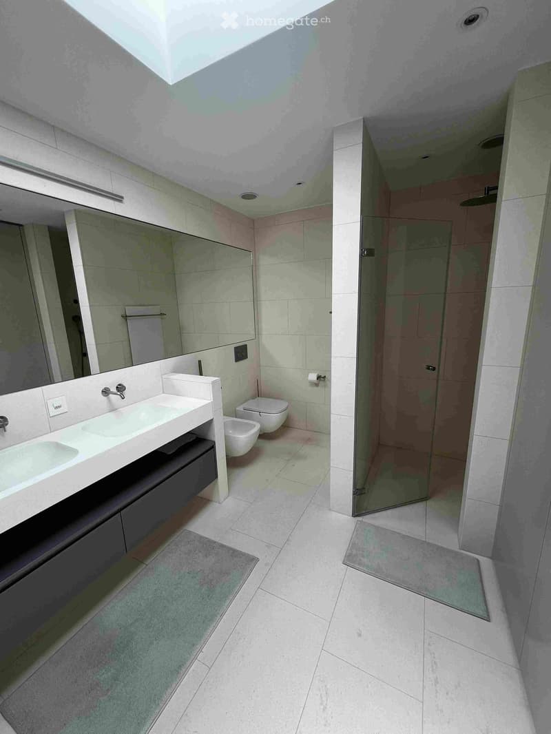 Modern bathroom with a double sink vanity, large mirror, toilet, bidet, and a walk-in shower with glass enclosure. The space features light-colored tiles and a skylight for natural light.