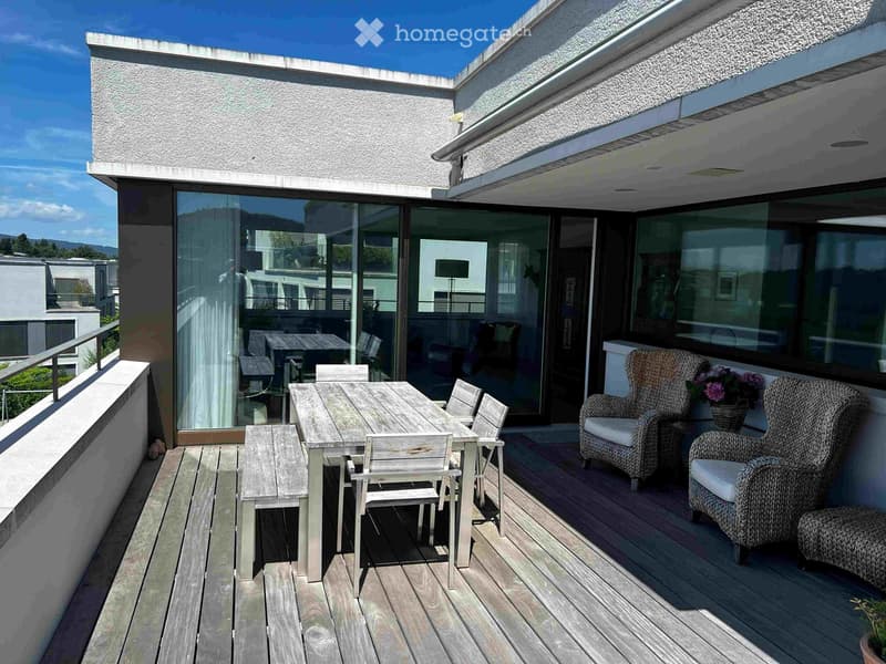 Wooden deck with a dining table and chairs, wicker armchairs, and a flower pot, with large glass doors leading to the interior.