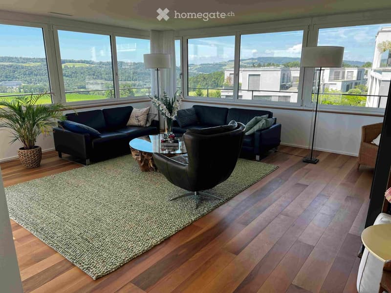 Modern living room with large windows offering a scenic view, featuring a black sectional sofa, a glass coffee table, a leather armchair, a floor lamp, a green rug, and a potted plant.