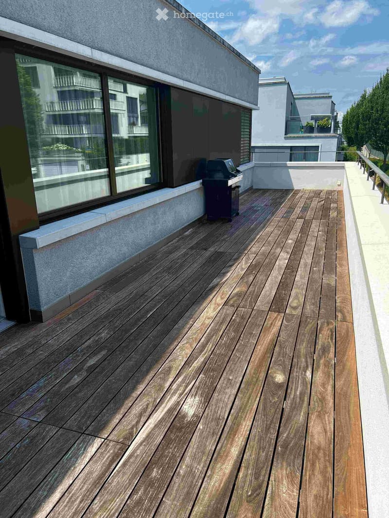 Wooden decked balcony with a grill, large windows, and a view of neighboring buildings.