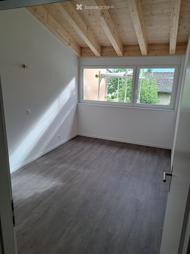 Empty room with wooden flooring, white walls, and a large window with a view of the outside. The ceiling has exposed wooden beams.