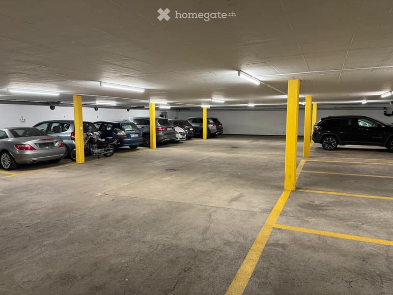 Underground parking area with several parked cars, including sedans, SUVs, and a motorcycle, separated by yellow-painted pillars and marked parking spaces.