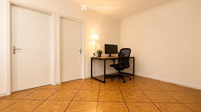 A small, well-lit room with wooden flooring, a desk with a computer monitor, a chair, a lamp, and a small plant. Two white doors are visible on the left side.