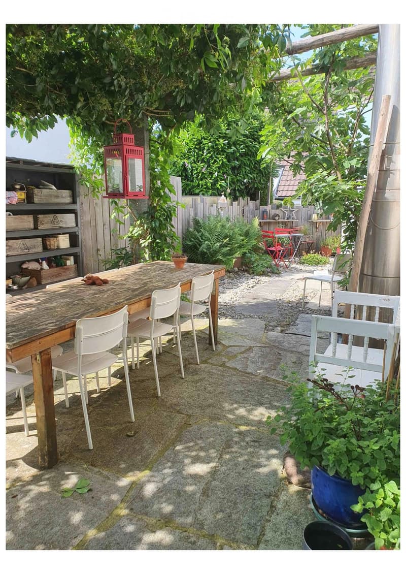 A cozy outdoor garden area with a wooden table, white chairs, potted plants, and a lantern hanging from a tree branch.