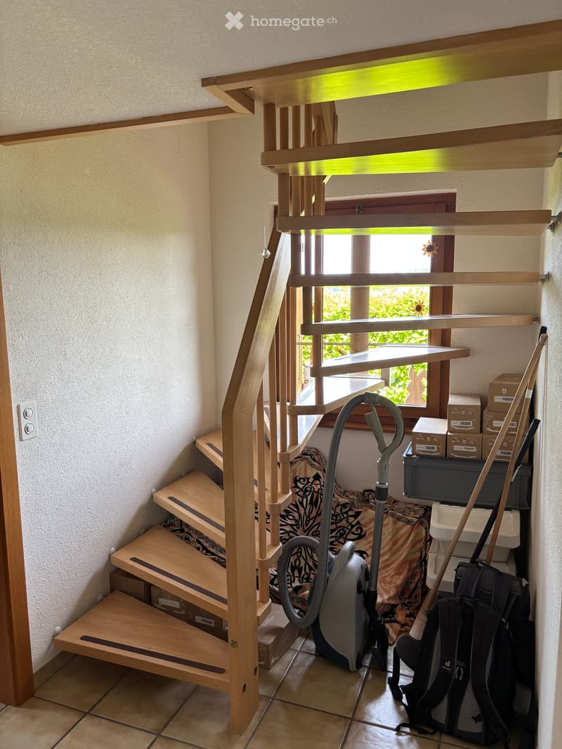 Wooden staircase with handrails and steps, adjacent to a window showing greenery outside. Cleaning supplies and storage boxes are placed near the stairs.