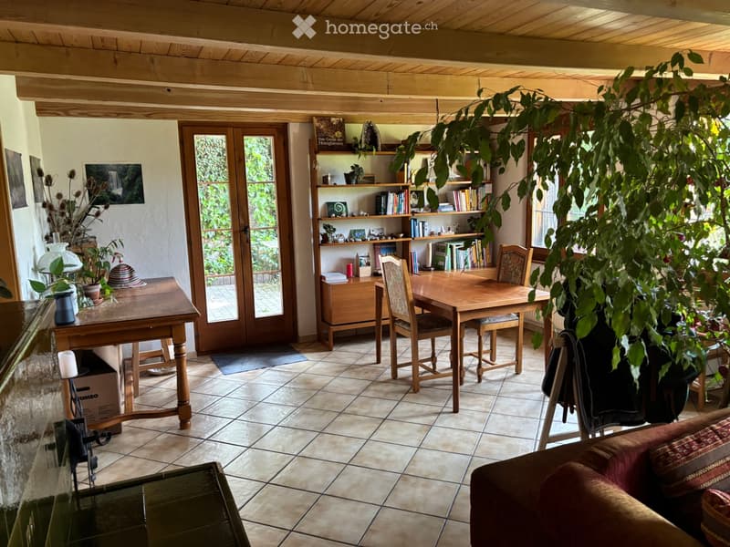 A cozy, well-furnished room with wooden furniture, including a sofa, dining table, chairs, bookshelf, and plants. The room features large windows and a wooden ceiling.